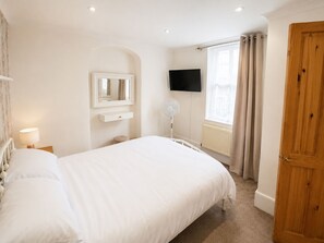 A bedroom with a bed, a bedside table, a mirror, a TV, a window with curtains, and a wooden door. - Muckoroos Cottage | Historic Flint Cottage (Ramsgate)
