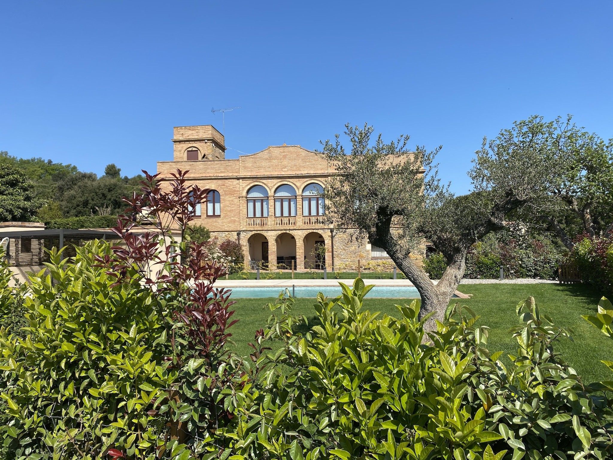 Un edificio grande con múltiples arcos y una torre, rodeado de jardines cuidados y una piscina.