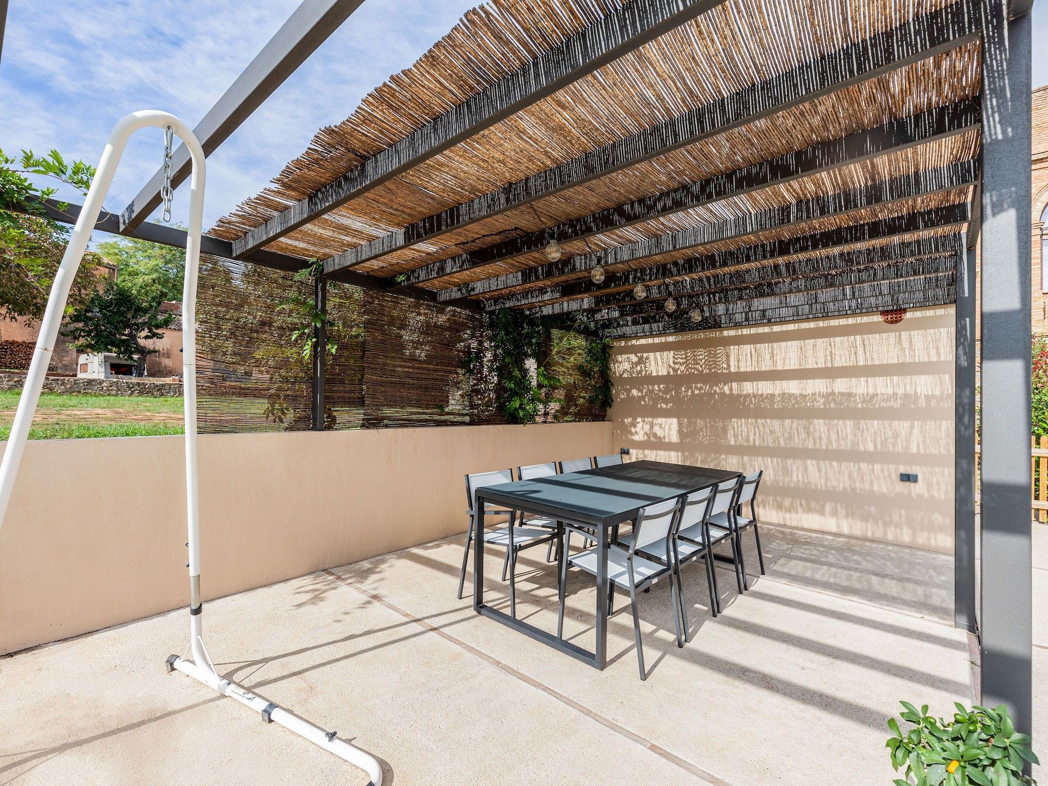 An outdoor patio with a table and chairs, a pergola with a woven roof, and a hose reel.