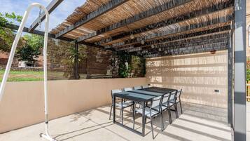 An outdoor patio with a table and chairs, a pergola with a woven roof, and a hose reel.