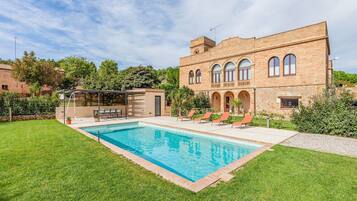 Una piscina con zona ajardinada, un porche cubierto y un edificio con ventanas de arco y una torre.