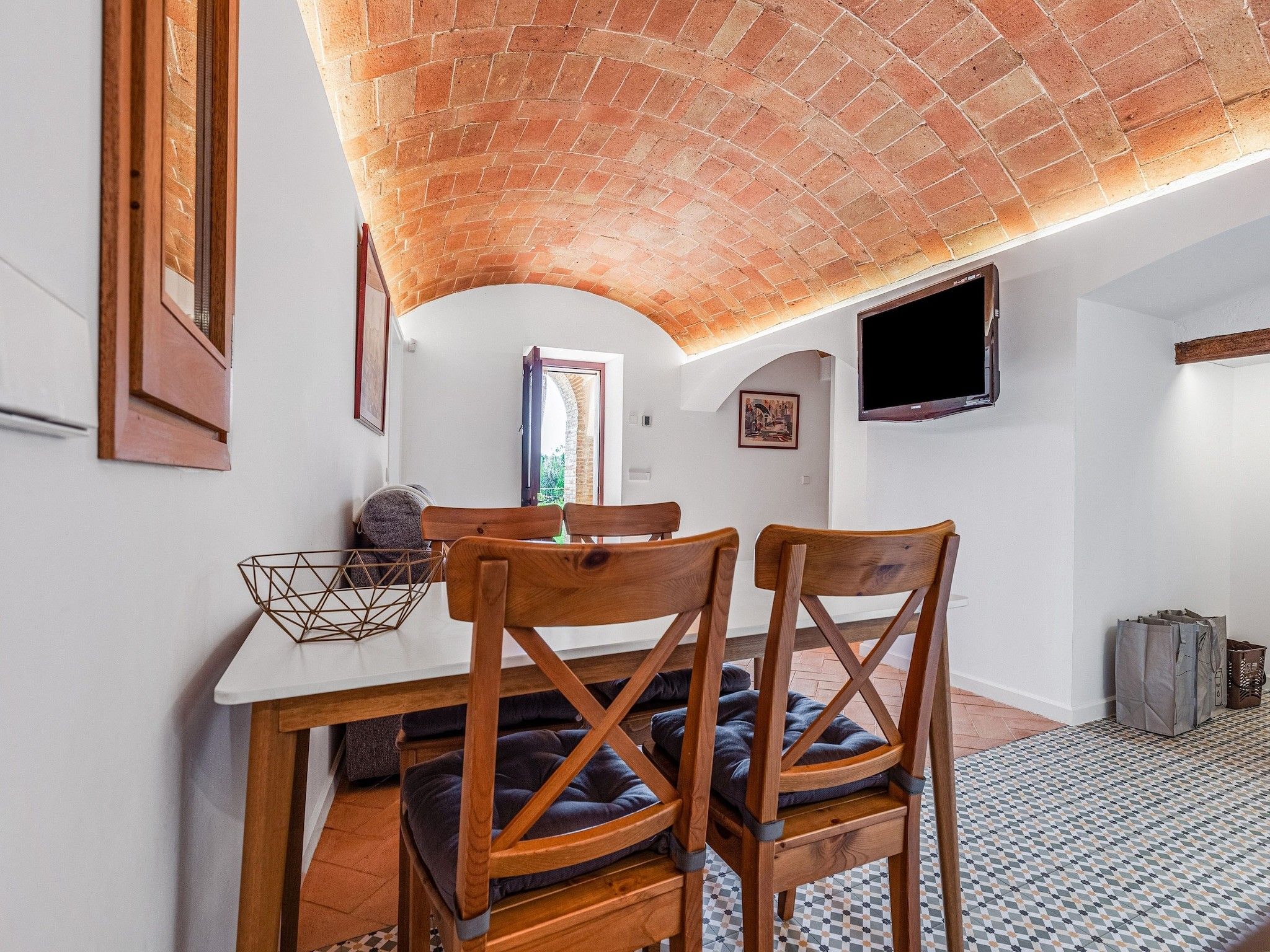 A dining area with a brick ceiling, a table, two wooden chairs, a television, and a patterned floor.