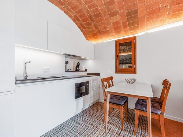 Une cuisine compacte avec des meubles blancs, un four encastré et un coin repas comprenant une table blanche et des chaises en bois.