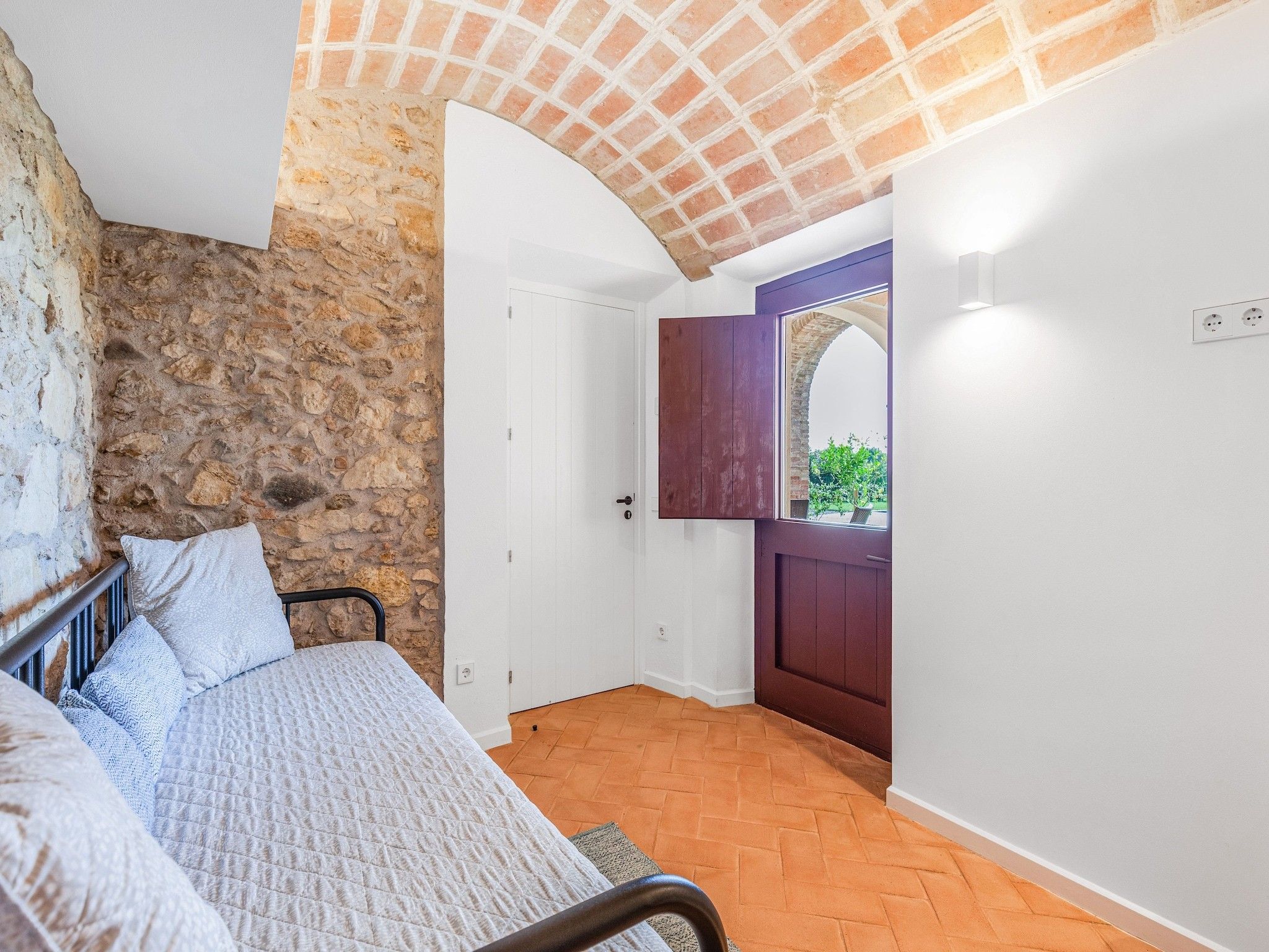 Un dormitorio con pared de piedra, una cama con colcha, una puerta de madera y vistas a través de un arco.