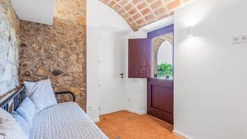 Un dormitorio con pared de piedra, una cama con colcha, una puerta de madera y vistas a través de un arco.