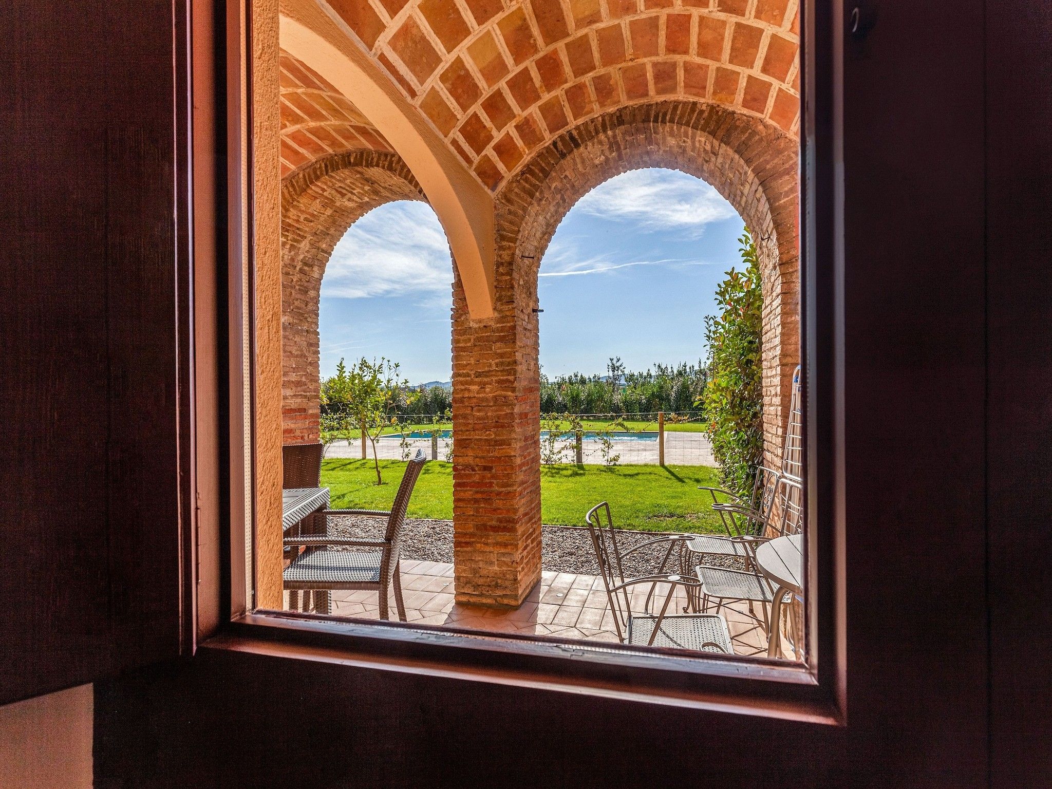 Visão pela janela, com um arco de tijolos que dá para um pátio externo com cadeiras e mesas.