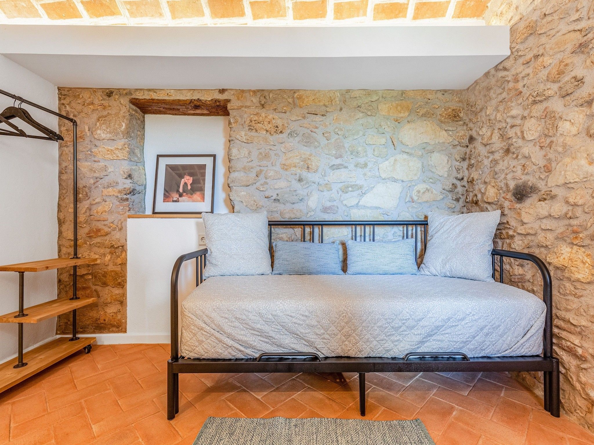 Een slaapkamer met een stenen muur, een bed met metalen frame, een houten plank en een foto aan de muur.