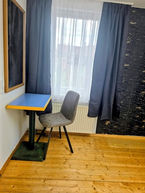 A small room with a wooden floor, a blue table, a grey chair, and a window with white curtains. - Vollmöbliertes 4-Zimmer-Haus in Pforzheim (Pforzheim)