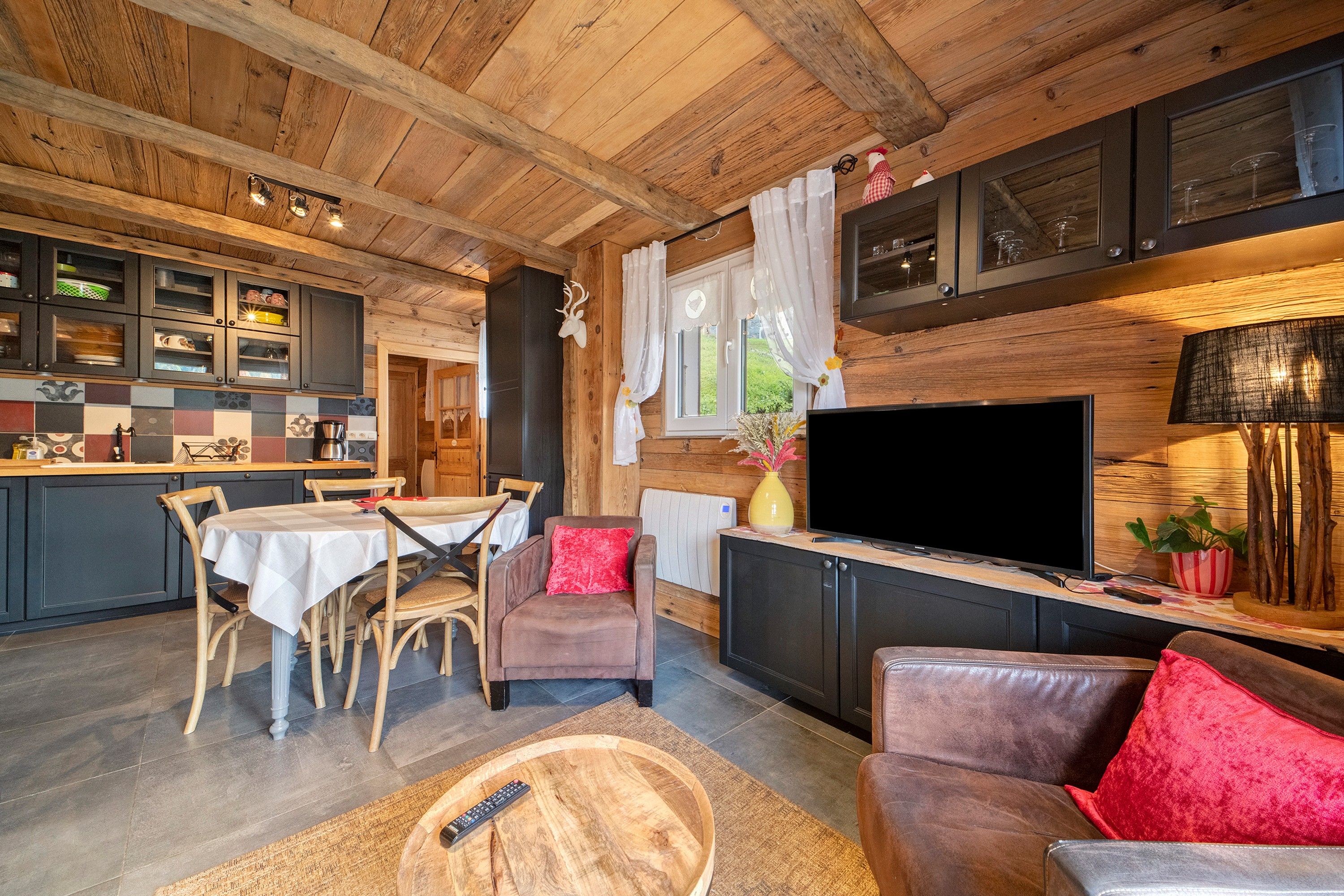 Een gezellig chaletinterieur met een eethoek, een woonkamer met een bank en televisie, en een keuken met houten kastjes en een groot raam.