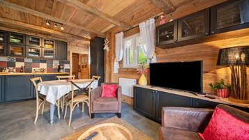 Een gezellig chaletinterieur met een eethoek, een woonkamer met een bank en televisie, en een keuken met houten kastjes en een groot raam.