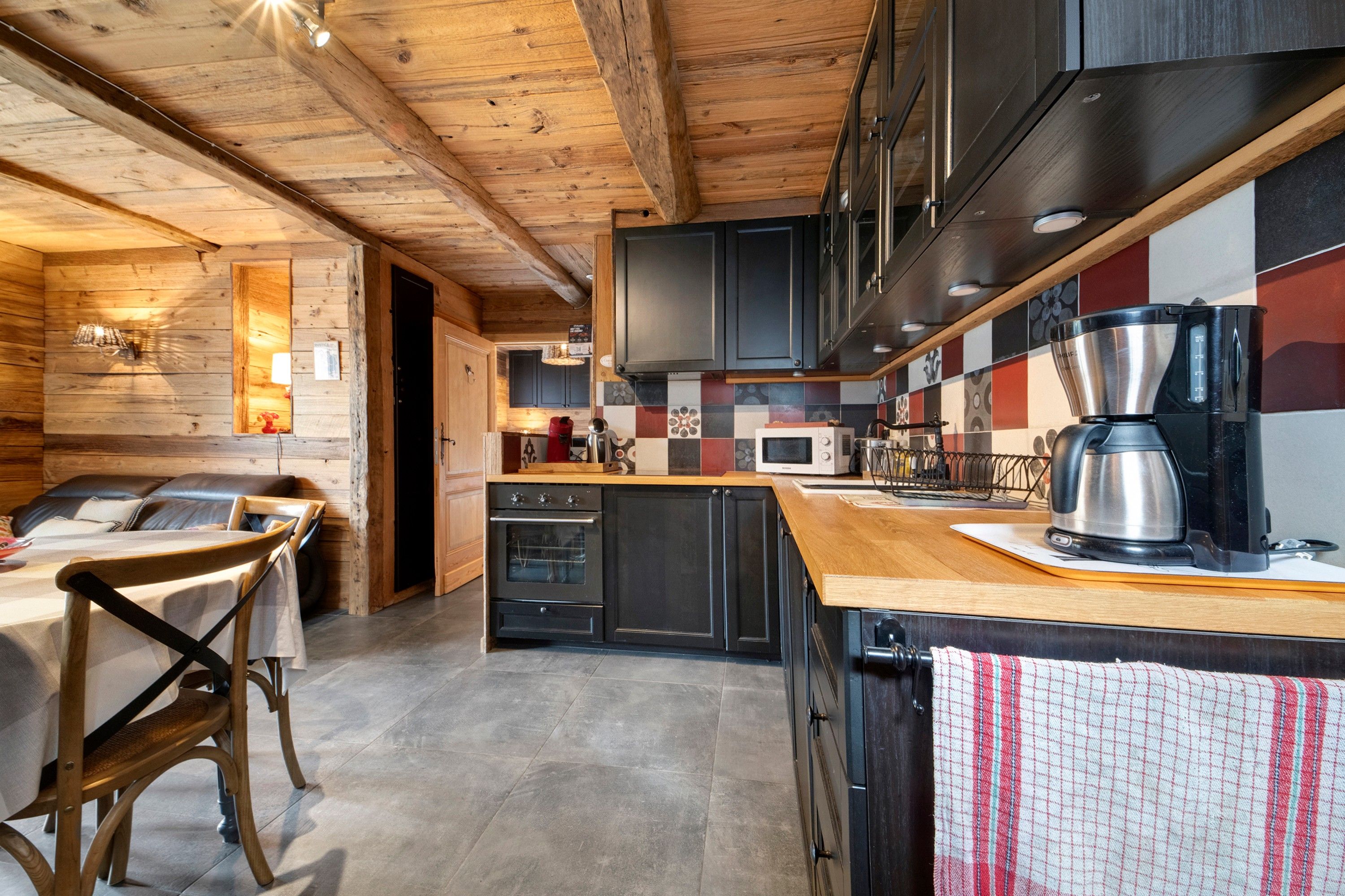 A rustic kitchen with wooden beams, a dining area with a table and chairs, and a tiled backsplash.
