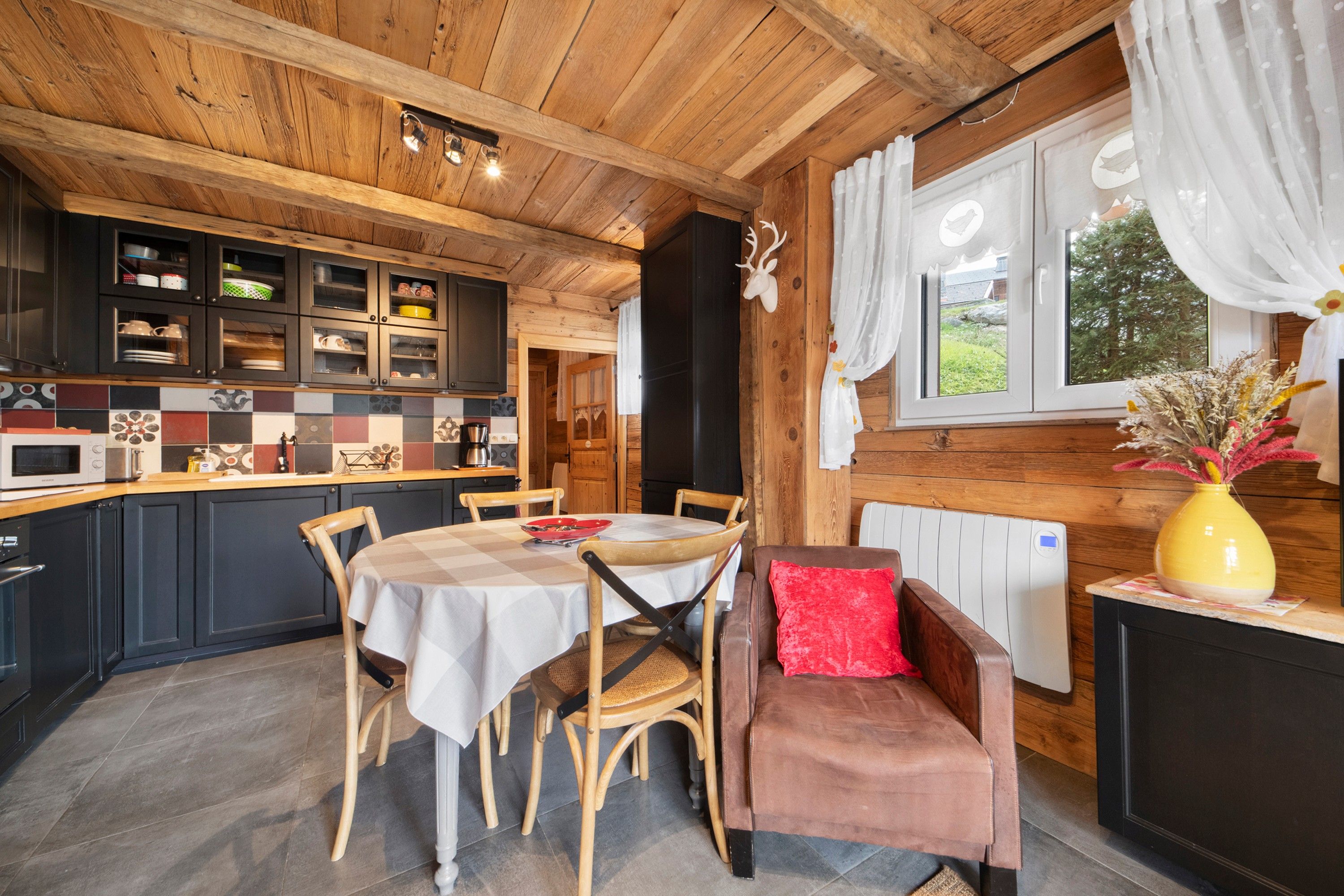 A cozy cabin interior with a dining table, chairs, a sofa, and a kitchen area with cabinets and a window.