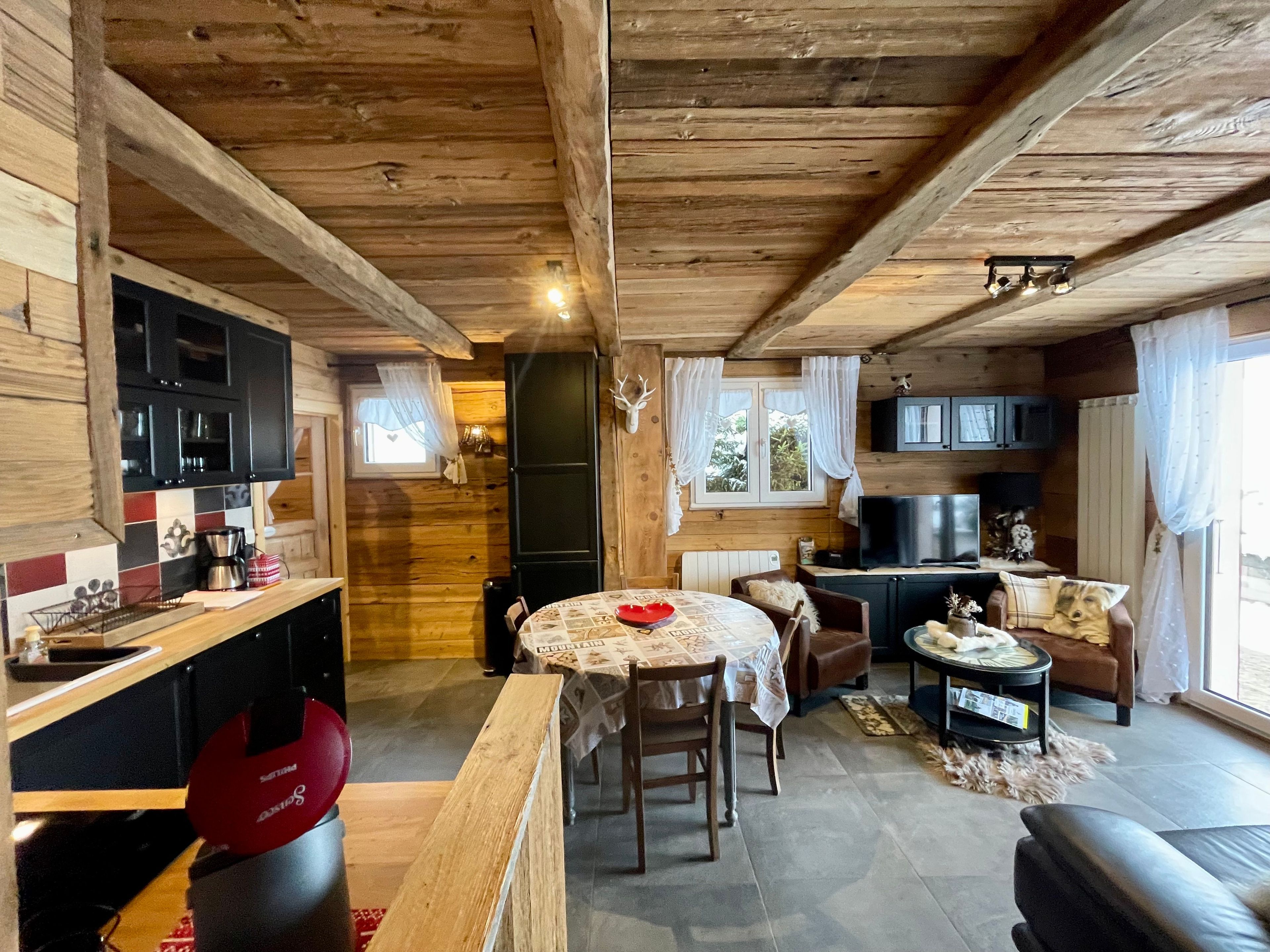 L’intérieur chaleureux d’un chalet avec un plafond en bois, un coin repas comprenant une table et des chaises, une cuisine équipée de placards et d’une cuisinière, et un salon avec un canapé et un téléviseur.