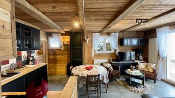 L’intérieur chaleureux d’un chalet avec un plafond en bois, un coin repas comprenant une table et des chaises, une cuisine équipée de placards et d’une cuisinière, et un salon avec un canapé et un téléviseur.