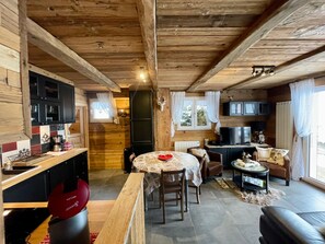 L’intérieur chaleureux d’un chalet avec un plafond en bois, un coin repas comprenant une table et des chaises, une cuisine équipée de placards et d’une cuisinière, et un salon avec un canapé et un téléviseur.