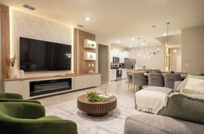 A modern living room with a flat-screen TV, green sofas, a coffee table, and a kitchen in the background. - 7BR house sleeps 20 with game room and pool (Clermont)