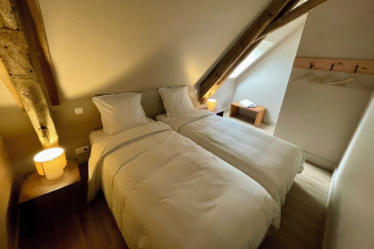 Un dormitorio con techo inclinado, una cama con ropa de cama blanca, una lámpara de noche y una mesita de noche de madera.