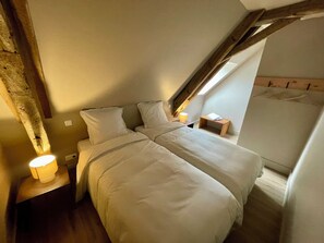 Un dormitorio con techo inclinado, una cama con ropa de cama blanca, una lámpara de noche y una mesita de noche de madera.