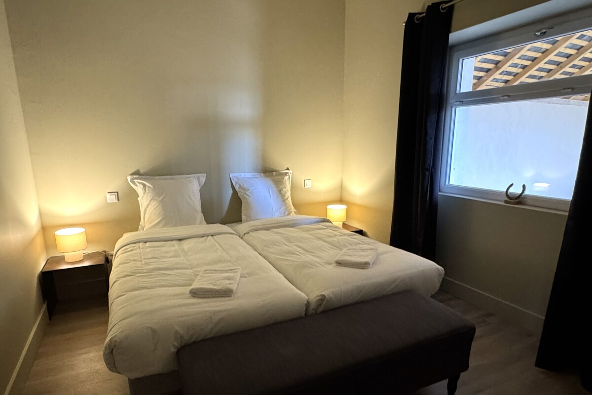 Una habitación de hotel con una cama, dos lámparas de noche, un ventanal con cortinas y suelo de madera.