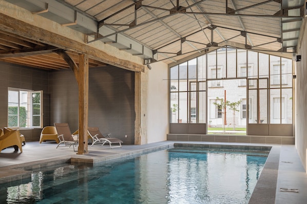 An indoor swimming pool with clear water, surrounded by lounge chairs and a high ceiling with a metal framework.