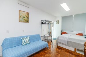 A room with a blue sofa, a bed with a white bedspread, a wooden floor, a wall-mounted mirror, and a painting. - Studio in Ipanema • Next to the Beach and Lagoon (Rio de Janeiro)