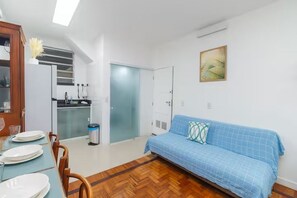 Un espace de vie compact comprenant un coin cuisine, un coin repas avec une table dressée pour deux personnes, et un canapé bleu placé contre le mur.