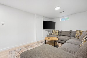 Ein modernes Wohnzimmer mit einem grauen Sofa, gemusterten Kissen, einem kleinen Holztisch und einem an der Wand befestigten Fernseher.