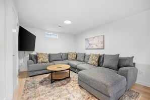 A modern living room with a grey sectional sofa, a round coffee table, patterned throw pillows, a large flat-screen TV mounted on the wall, and a patterned rug on the floor. - Falmouth Group Retreat, Outdoor Living, Central AC, Dogs OK (Falmouth)