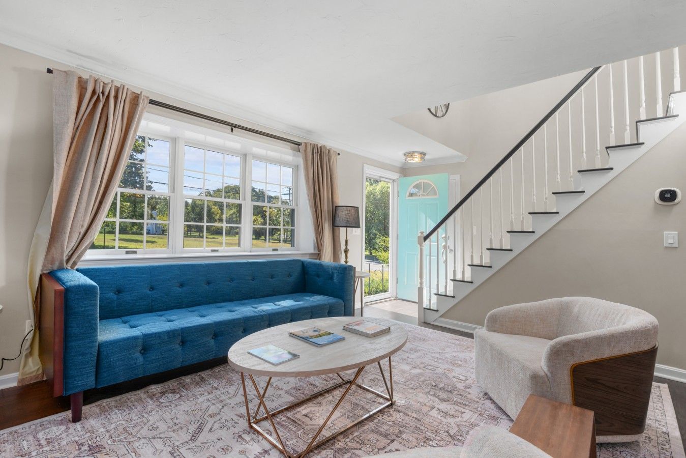 Ein Wohnzimmer mit einem blauen Sofa, einem runden Couchtisch, einem hellen Sessel, einer Treppe und Blick ins Freie.