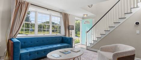 A living room with a blue sofa, a round coffee table, a light-colored armchair, a staircase, and a view of the outdoors.