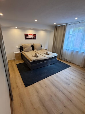 A modern bedroom with a large bed, a nightstand, a wall-mounted picture, and a window with curtains.