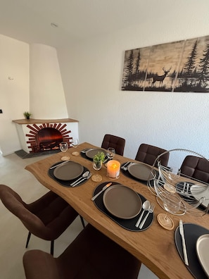 A dining area with a wooden table set for six, surrounded by chairs, with a fireplace and a forest-themed painting on the wall.