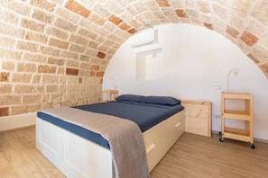 A bedroom with a brick wall, a bed with a blue mattress, a wooden nightstand, a bedside table, and a wall-mounted air conditioning unit. - Torretta Morelli by BarbarHouse (Sannicola)