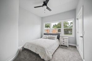 A bedroom with a bed, a nightstand, a dresser, a ceiling fan, and a window with a view of a house. - Dreamy Duplex B-Spacious & Comfortable! 3 Bedrooms! (Hartford)