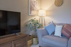 Ein Wohnzimmer mit einem Flachbildfernseher auf einem Holzboden, einem grauen Sofa mit Kissen, einer Stehlampe und einer Topfpflanze.