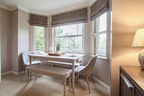 Ein Essbereich mit Tisch, Stühlen und einer Bank. Es gibt Fenster mit Vorhängen, eine Lampe und eine Pflanze auf dem Tisch.