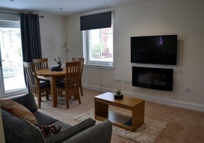 Una sala de estar con un sofá, una mesa de comedor con sillas, un televisor montado en la pared y una pequeña planta sobre una mesita auxiliar.
