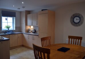 A modern kitchen with wooden cabinets, a round dining table, and a wall clock. - 6 Cara Anan- 2 Bedroom House (Stirling)