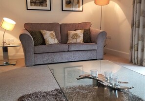 A grey sofa with patterned cushions, a glass coffee table with a metal tray holding glasses, and a lamp on a side table. - 12 Alainn Eve - 2 Bedroom House, Killin (Stirling)