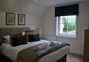 A bedroom with a bed, pillows, a blanket, a lamp, a window with a view, and two framed pictures on the wall. - 7 Gleann Fia House - 2 Bedroom House Killin (Stirling)