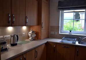 A kitchen with wooden cabinets, a kettle, a toaster, and a window with a view of greenery. - 7 Gleann Fia House - 2 Bedroom House Killin (Stirling)