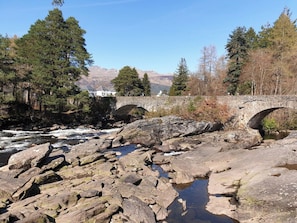 A rocky riverbank with a stone bridge and a house in the background. - 7 Gleann Fia House - 2 Bedroom House Killin (Stirling)
