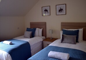 Ett hotellrum med två sängar, var och en med ett blått, vadderat sängöverkast och vita sängkläder. Ovanför sängarna hänger inramade tavlor på väggen.