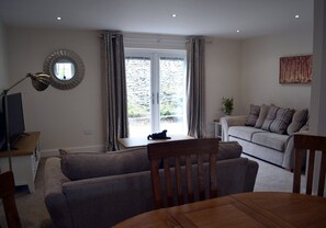 A living room with a sofa, a dining table, a TV, and a window with curtains. - 3 Dorcha's Mian - 2 Bedroom House, Killin (Stirling)