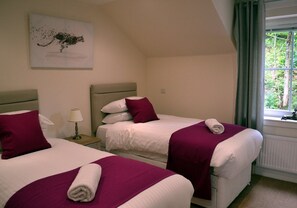 Quarto de hotel com duas camas, cada uma com cabeceira branca, colcha roxa e toalha branca enrolada. Há uma janela com cortinas, um radiador e um quadro na parede.