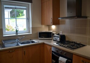 A kitchen with a gas stove, microwave, toaster, sink, and wooden cabinets. - 9 Inverfinn - 2 Bedroom House Killin (Stirling)