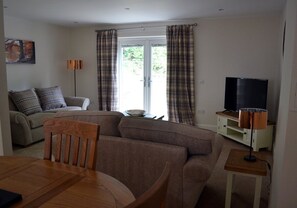 A living room with a sofa, a dining table with chairs, a television, and curtains on a window. - 9 Inverfinn - 2 Bedroom House Killin (Stirling)