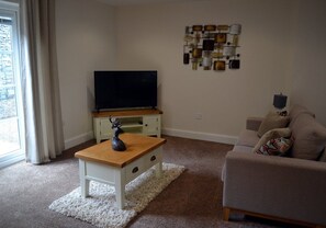 A living room with a flat-screen TV on a stand, a white coffee table with a decorative item, a beige sofa, and a wall-mounted light fixture. - Orangewood House (4) - 2 Bedroom House, Killin (Stirling)