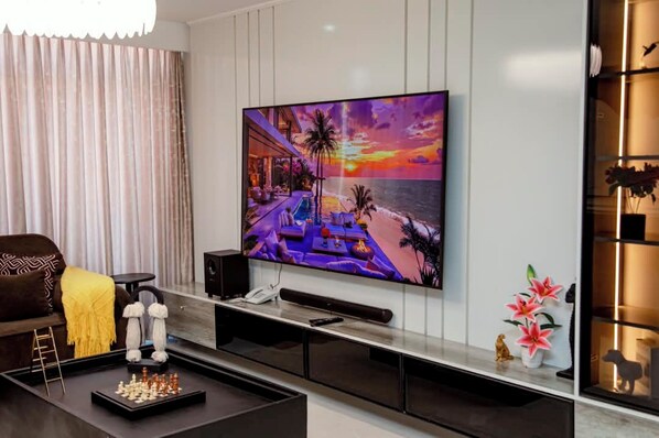 A modern living room with a flat-screen TV mounted on the wall, a coffee table with a chess set, a sofa, and a decorative shelf with plants. - 5 minutes to the theme park! Family-sized condo with game room & deck (Dar es Salaam)