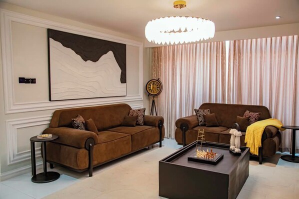 A modern living room with two brown sofas, a coffee table with chess pieces, a large abstract painting, a clock, and a chandelier.